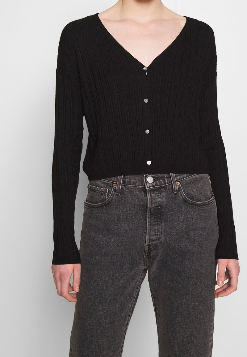 Personne portant un cardigan noir à boutons à col en V côtelé et un jean noir délavé taille haute, debout devant un fond uni.