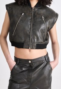 Przycięta kamizelka ze skóry w ciemnym brązie z srebrnymi zamkami błyskawicznymi, nakładkami na ramiona i prążkowanym pasem. W zestawie z dopasowaną skórzaną spódnicą.