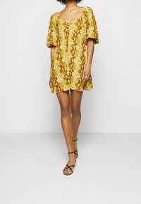 Gul kort klänning med puffärmar, prydd med en texturerad blommönster. Bärs med svarta sandaler med remmar, stående mot en vit bakgrund.