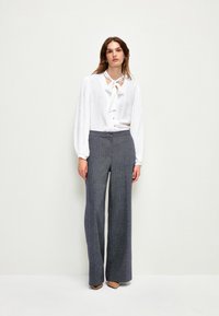 Vit blus med nyckelhålsutskärning och rosettdetalj, kombinerad med högmidjade, vida grå byxor med textur och neutrala klackskor.