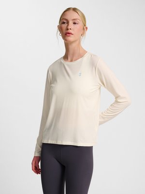 Mujer con cabello rubio que lleva una camisa de manga larga crema y leggings negros, de pie con una mano en la cadera sobre un fondo liso.