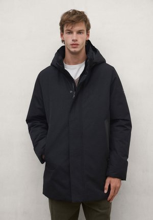 Young man wearing a black hooded winter coat with hands in pockets standing against a plain light background.