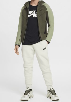 Mandlig model iført en tofarvet grøn Nike-hoodie med lynlås, sort "Just Do It" Nike t-shirt, hvide Nike træningsbukser og hvide Nike sneakers.