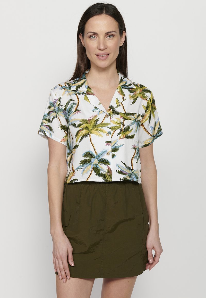 Gekürzte weiße Bluse mit grünem und gelbem Palmenmuster, kurzen Ärmeln und durchgehender Knopfleiste, kombiniert mit einem olivgrünen Rock.