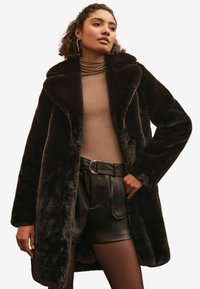 Femme portant un manteau en fausse fourrure noire, un col roulé beige, un short en cuir noir ceinturé et des collants noirs sur fond uni.