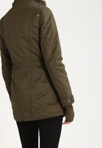 Osoba oblečená v olivově zelené přiléhavé bundě s knoflíkovými manžetami a černými kalhotami, zobrazená zezadu na bílém pozadí.