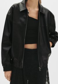 Czarna kurtka bomberka z faux skóry z kołnierzykiem, zamkiem błyskawicznym z przodu, elastycznymi mankietami i gładką fakturą. Akcentowana eleganckim designem.