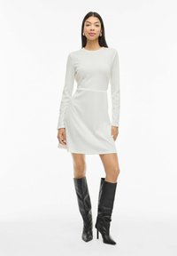 Robe à manches longues côtelée de couleur gris clair, cintrée à la taille. Associée à des bottes stilettos noires montant jusqu'aux genoux. Texture lisse, design simple.