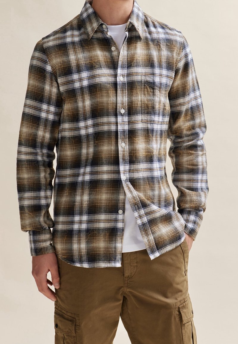 Homme portant une chemise à carreaux marron et blanche boutonnée sur un t-shirt blanc avec un pantalon cargo marron devant un fond neutre.