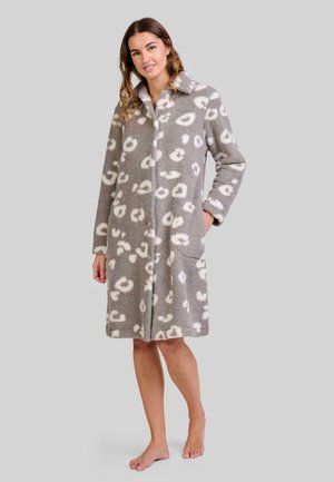 Mujer de pie descalza, vistiendo una bata de felpa gris con manchas de leopardo blancas y bolsillos, sonriendo frente a un fondo sencillo.