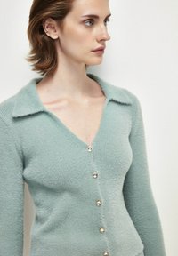 Ljusgrön cardigan gjord av textil, mjukt material. Har V-ringning, långa ärmar och fem dekorativa knappar fram.