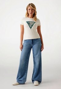 Valge lühikeste varrukatega T-särk rohelise ja musta kolmnurga kujundusega esiküljel, kombineeritud helesiniste laiade jalgadega teksapükste ja valgete tossudega.