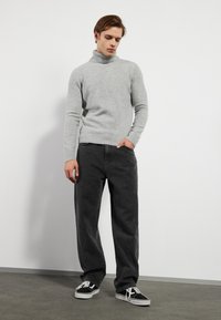 Lys grå turtleneck genser i myk stoff, parret med svarte vide jeans. Modellen har på seg svart-hvite joggesko og står mot en ensfarget bakgrunn.