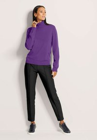 Frau in lila Rollkragenpullover, schwarzen maßgeschneiderten Hosen und dunklen Sneakern, die an einer hellen Wand steht und zur Seite schaut.