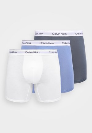 Tre boxer da uomo Calvin Klein in bianco, azzurro chiaro e grigio scuro con cinturini bianchi con logo, impilati uno accanto all'altro.