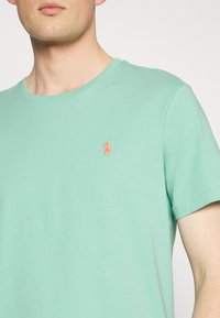 Mentolově zelené tričko s krátkým rukávem, kulatým výstřihem, vyrobené z měkké látky, s vyšívaným oranžovým logem na levé části hrudníku.