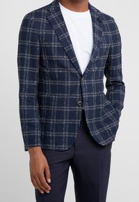 Marinblå kavaj med ett rutmönster i vitt och ljusblått. Har en tvåknappsstängning, slagkragar och textil med struktur.