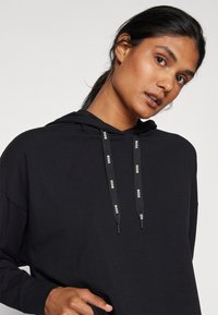 Felpa nera con cappuccio realizzata in tessuto morbido, caratterizzata da un cordino con dettagli del logo e una vestibilità rilassata. Maniche lunghe e cappuccio completano il design.