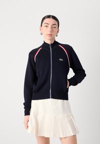 Jūrų laivyno zip-up džemperis su aukštu apykakle, raudonomis ir baltomis juostelėmis ant pečių bei žaliu Lacoste logotipu kairėje krūtinės pusėje.