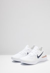 White athletic shoes with a textured knit upper, laces, and a black swoosh logo. Features a cushioned sole with a light brown accent.