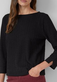 Vrouw in een zwart gestructureerd langslijf met een kleine openingssleeve, hand in de zak, gekleed in bordeauxrode broek tegen een grijze achtergrond.