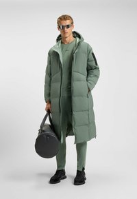 Grüne Daunenjacke mit Kapuze, kombiniert mit passender Sportbekleidung. Enthält eine schwarze Sporttasche und sportliche Schuhe mit strukturierten Sohlen.