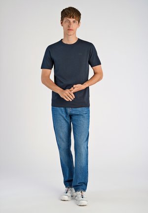 Camiseta de algodón azul marino con mangas cortas y cuello redondo, combinada con jeans de denim azul claro. El modelo está de pie con las manos entrelazadas.
