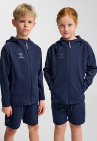 Två barn bär matchande marinblå hujackor. Jackorna har blixtlåsförslutning, sidofickor och grå detaljer på axlar och ärmslut.