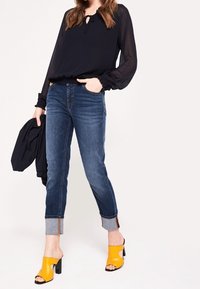 Camicia nera con colletto a fiocco, jeans blu con orli risvoltati e mule gialle con tacco a blocco. L'outfit è completato da una giacca nera.