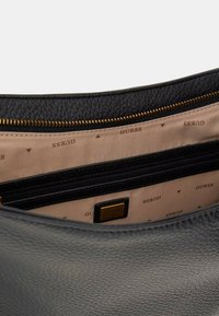 Zwarte leren handtas met een gestructureerde buitenkant, gouden rits en een beige interieur met herhaaldelijk "GUESS"-logo en een ritszak.