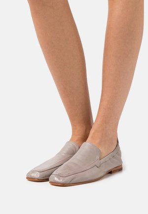 Fødder iført lys grå læder slip-on loafers med firkantede tæer og flade træsåler på en hvid baggrund.