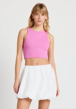 Giovane donna con top corto rosa senza maniche e gonna bianca a palloncino, in piedi davanti a uno sfondo chiaro uniforme.