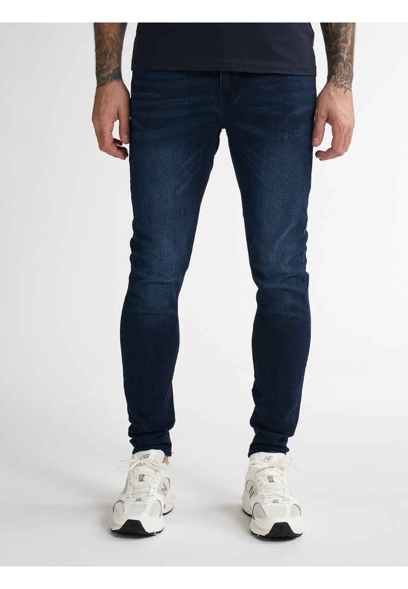 Tmavě modré džíny slim fit vyrobené z denimu, s jemnou strukturou a designem s pěti kapsami, doplněné bílými sportovními botami.