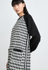 Musta-valkoinen houndstooth-kuosiset mekko, jossa on raglan-hihat, mustat taskut ja sivuraidat. Teksturoitu kangas ja rento leikkaus.