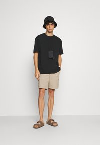 Černé oversized tričko, béžové cargo šortky, hnědé sandály. Doplňky: malá černá taštička na popruhu. Jednoduchý design, casual outfit.