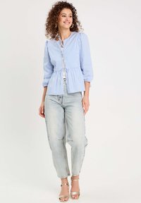 Blusa de botones a rayas azul y blanca con cintura peplum, mangas tres cuartos y cierre de lazo en el cuello, combinada con jeans claros y sandalias.