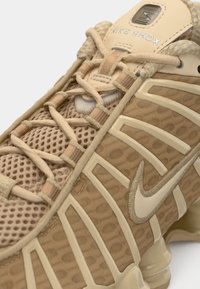 Μπεζ αθλητικό παπούτσι Nike Shox με επάνω μέρος από υφασμάτινο πλέγμα με υφή και λεία συνθετικά επικαλύμματα. Διαθέτει εμφανές σύστημα κορδονιών και επενδεδυμένη σόλα.