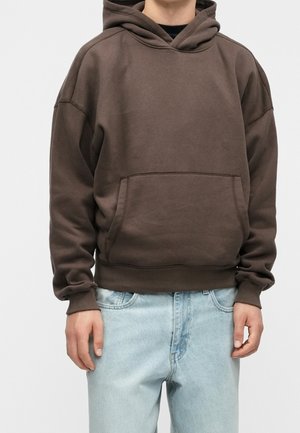 Person iført en mørkebrun hættetrøje med forlomme og lyseblå jeans, stående mod en ensfarvet baggrund.