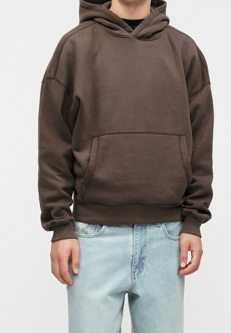 Personne portant un sweat à capuche marron foncé avec une poche avant et un jean bleu clair, debout devant un fond uni.