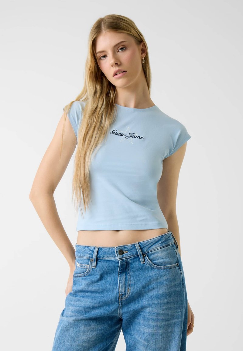 Lichtblauw crop top van zachte katoen, met korte mouwen en zwarte "Guess Jeans" borduursel boven een sterontwerp. Draagt een blauwe spijkerbroek.