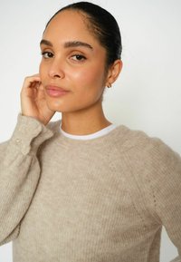 Bežani pleteni pulover, izrađen od mekanog prediva s teksturiranim završetkom. Ima okrugli izrez i gumbe kao detalj na rukavu.