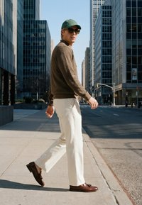 Homme portant une casquette verte, des lunettes de soleil, un pull marron, un pantalon blanc et des chaussures marron, marchant sur le trottoir de la ville entre de grands immeubles.