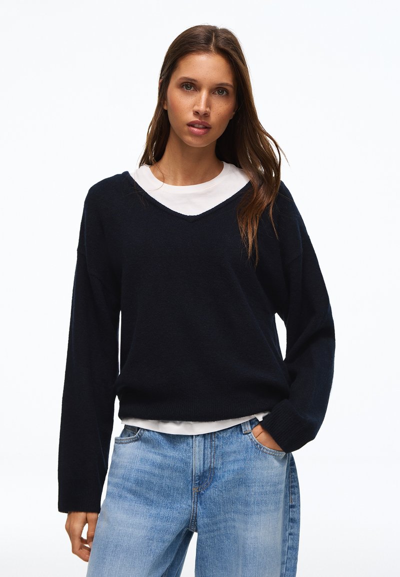 Femme aux longs cheveux bruns portant un pull noir à col en V par-dessus une chemise blanche et un jean bleu clair, une main dans la poche.