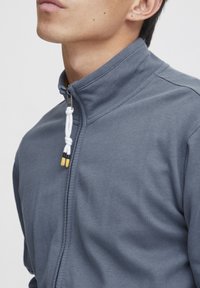 Grå zip-up jakke med høy krage, med kontrasterende hvit snor med gule og svarte detaljer. Myk bomullsblanding.