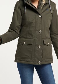 Kvinna iklädd en olivgrön jacka med huva och stora knäppta frampåsar samt blå jeans framför en enfärgad bakgrund.