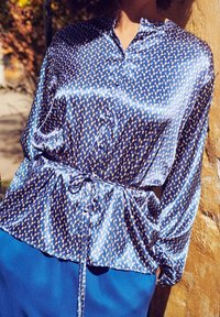 Modella in una blusa blu lucida a fantasia con vita legata, in piedi davanti a un muro texturizzato, con la luce del sole che illumina i suoi dintorni.