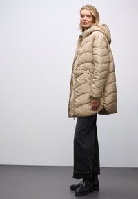 Beige puffjakke med hætte, quiltet mønster, lynlås lukning og sidelommer, parret med sorte brede jeans og sorte støvler.
