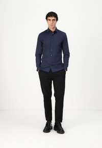 Mørkeblå langermet skjorte med knapper og slim fit, detaljert søm og spiss krage, kombinert med svarte bukser og sko.