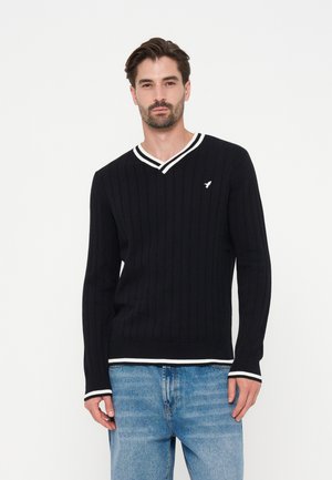 Man wearing black ribbed V-neck sweater with white trim and light blue jeans standing against plain white background.