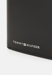 Crna kožna novčanica s utisnutim logotipom "TOMMY HILFIGER" u srebrnoj boji. Glatka tekstura s zaobljenim rubovima i vidljivim šavovima.